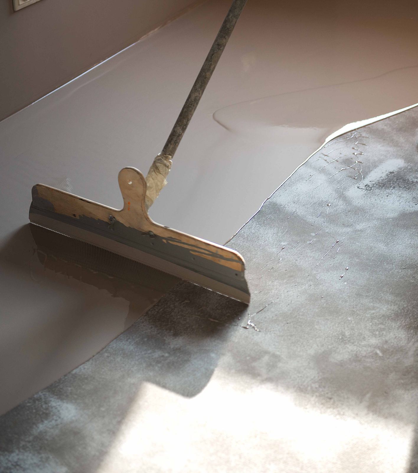 Squeegee spreading a light floor coating over gray concrete in a sunlit room